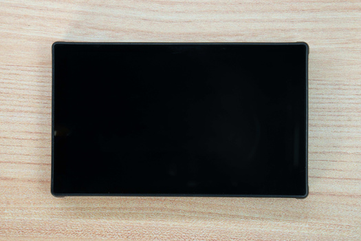 Black tablet or mobile device displaying a blank screen on light wood surface.