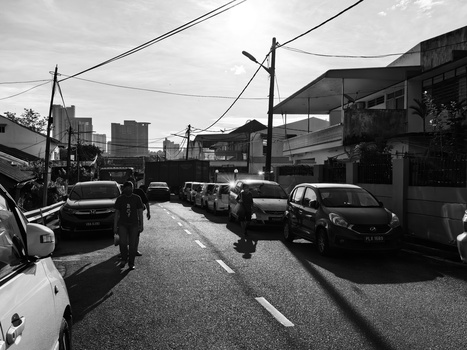 Black and white street scene with parked cars lined along an urban road beneath overhead power lines.