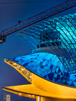 Modern architectural structure with blue geometric patterned facade and yellow curved element at dusk, illuminated by construction crane overhead.