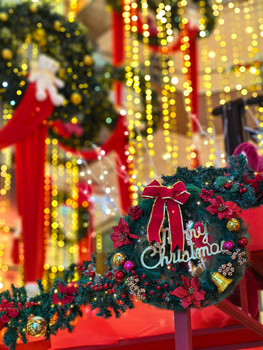 Close-up of a Christmas wreath with red bow and ornaments, with blurred holiday decorations in background.