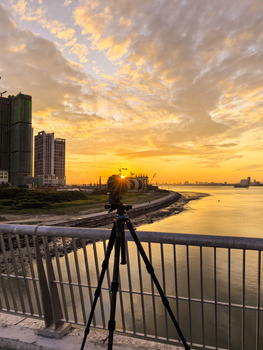 Camera on tripod mounted on bridge railing overlooking golden sunset over waterway and cityscape.