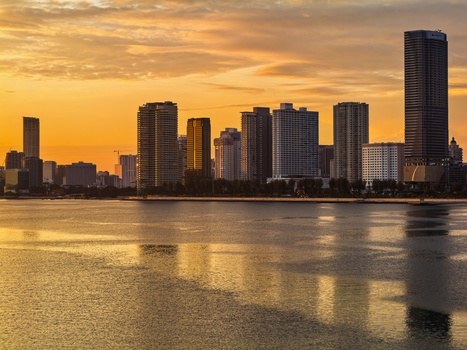 Urban skyline silhouetted against golden sunset sky, reflected in calm water below.