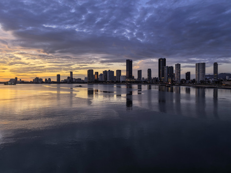 Urban skyline reflected in calm water at dawn, with purple and gold clouds overhead.