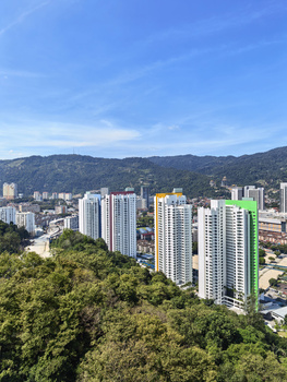 Aerial view of a dense urban residential district with high-rise apartment buildings nestled against forested hills under clear blue sky.
