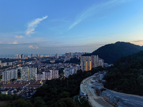 Cityscape at dusk with high-rise residential buildings, forested hillside, and curved highway in foreground.