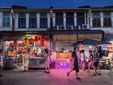 Night street scene with neon-lit market stalls and shoppers under colonial-era buildings.