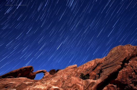 Star trails arc across a deep blue night sky above distinctive red rock formations with natural arches.