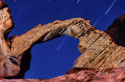 Natural stone arch formation silhouetted against deep blue twilight sky with star trails.