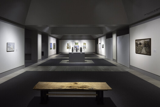 Contemporary art gallery with minimalist white and black walls displaying framed photographs and a wooden bench in foreground.
