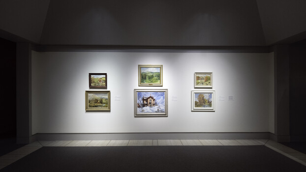 Gallery wall with six framed landscape paintings illuminated by museum-style spotlights against a white wall.