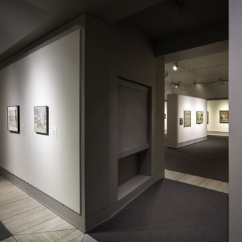 Modern gallery interior with framed artworks on white wall and open doorway leading to adjacent exhibition space.