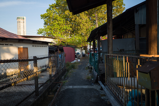Sunlit alley between residential buildings with chain-link fencing and mature trees overhead.