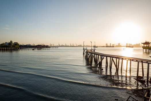 Wooden pier extending into calm bay waters at golden hour with distant cityscape and boats.