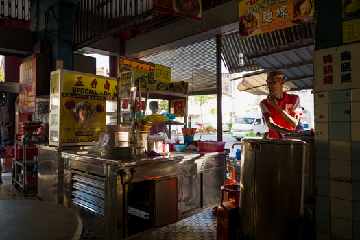 Man in red shirt working at a metal counter inside a cluttered Asian temple or shop with yellow signage and religious decorations.