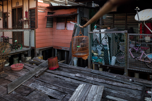 Weathered wooden deck extends toward a rusty metal house with cluttered patio storage and overgrown vegetation.
