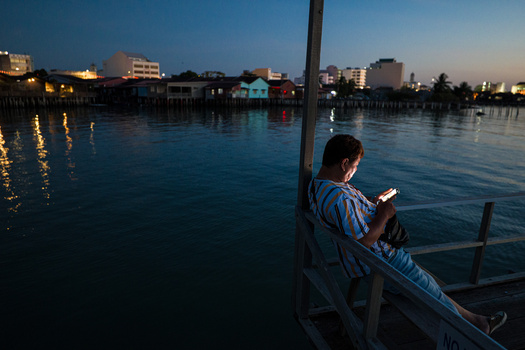 Man sitting on wooden boat at dusk, holding phone, overlooking waterfront buildings reflected in still water.
