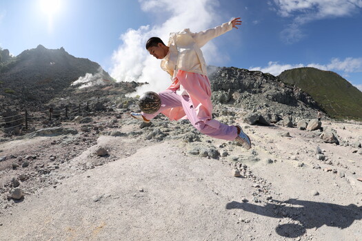 Skateboarder mid-trick on volcanic terrain with mountains and steam vents in background.