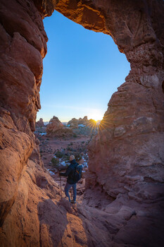 Hiker viewed from inside a narrow canyon passage, framed by red rock walls with blue sky and warm sunlight beyond.