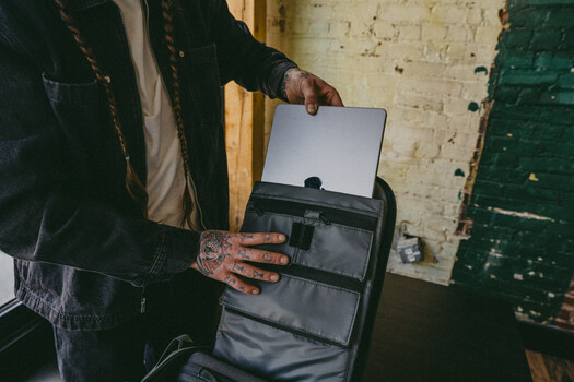 Person with tattooed hands holding an open camera bag against a weathered brick wall.