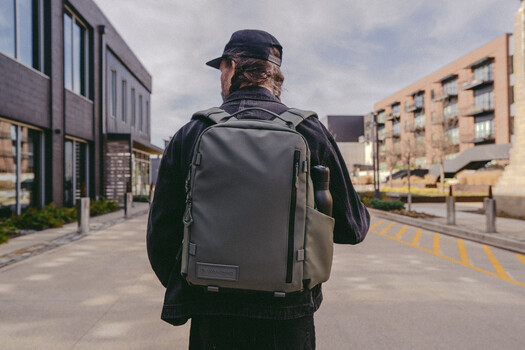Person wearing dark clothing and cap walks away from camera down urban street, carrying gray backpack.