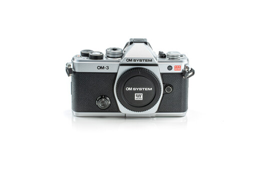 OM System OM-3 film camera body with chrome top plate and black textured grip, shown from front against white background.