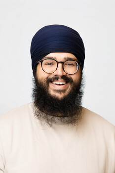 Man with full beard wearing glasses, navy beanie, and cream-colored shirt against neutral background.