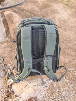 Gray camera backpack displayed from behind on rocky ground, showing padded straps and ventilated back panel.