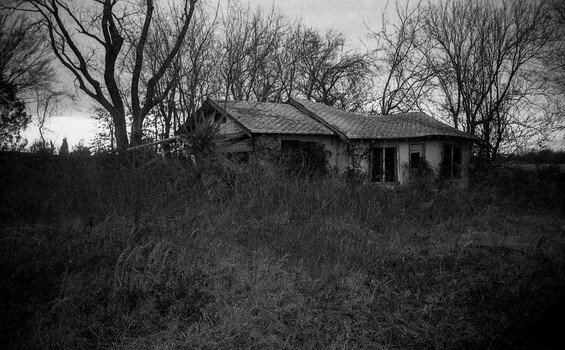 Abandoned single-story house surrounded by overgrown vegetation and bare trees in black and white.