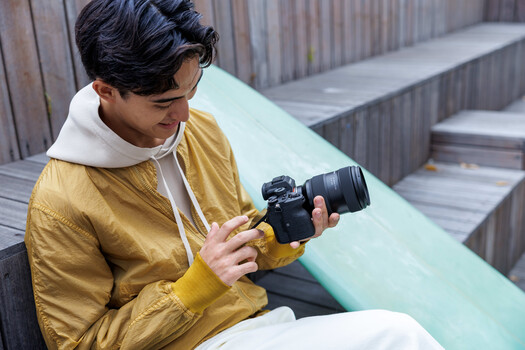 Photographer in yellow jacket examining a DSLR camera with attached telephoto lens on a modern outdoor bench.
