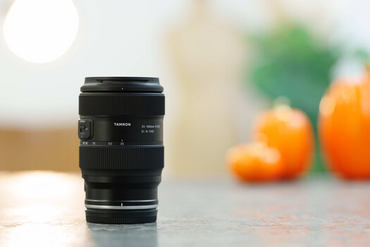 Tamron zoom lens positioned on a surface with blurred citrus fruits in the background.