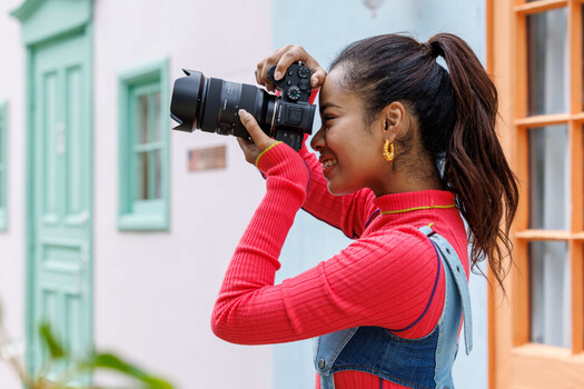 Young woman holding a DSLR camera with telephoto lens, photographing something off-camera while smiling.