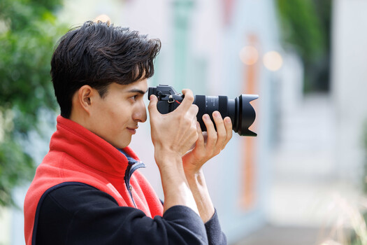 Young photographer in red jacket holding DSLR camera with telephoto lens to his eye.