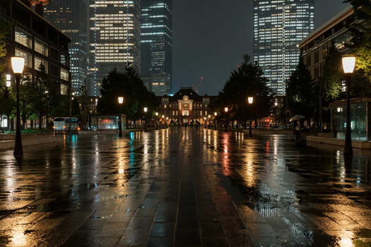 Wet urban plaza at night with illuminated skyscrapers and street lamps reflecting on pavement.