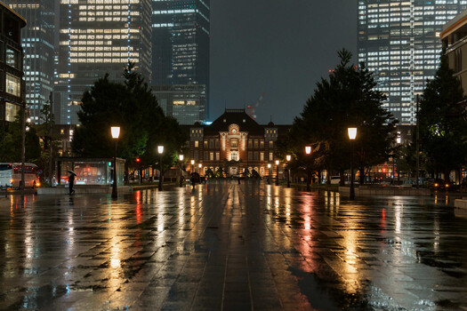 Wet plaza at dusk with illuminated historic building and modern skyscrapers reflecting on pavement.