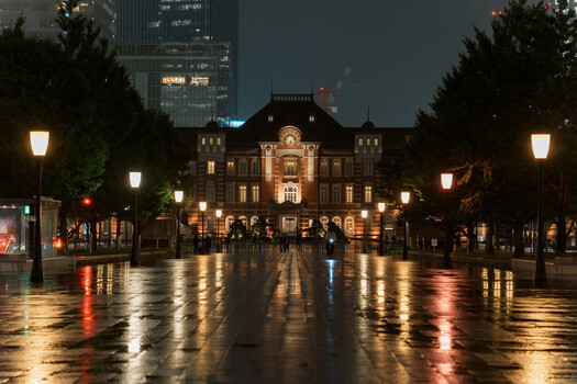 Wet plaza at dusk with illuminated historic building and street lamps reflecting on rain-soaked pavement.