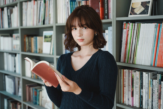 Woman holding an open book while standing in front of library shelves.