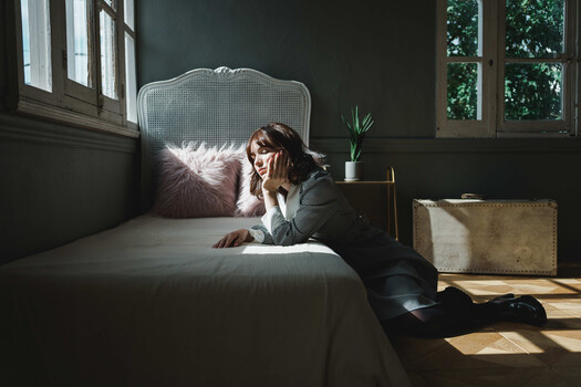 Woman in business attire sitting on bed near windows, hand to face in contemplative pose.