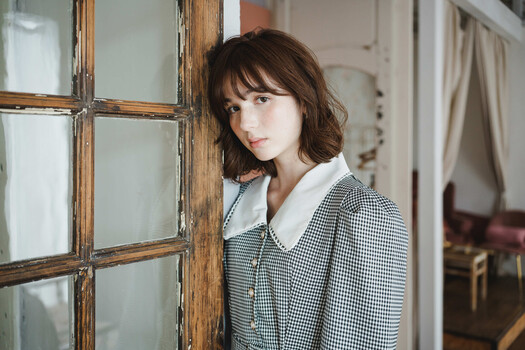 Woman in checkered blazer standing by wooden-framed window with diffused natural light.