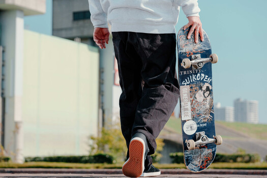 Skateboarder in white shirt and black pants holding a decorated skateboard deck with visible grip tape and graphics.