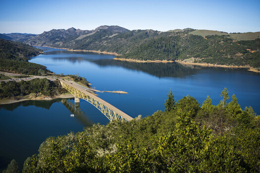 Aerial view of a blue reservoir with a concrete dam spanning between forested hillsides under clear sky.
