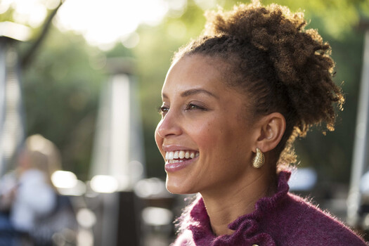 Woman with natural curly hair in an updo, smiling at the camera in profile against a blurred outdoor background.