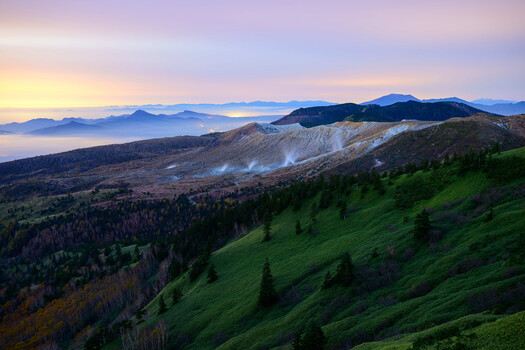 Misty valley with forested slopes beneath a dawn sky and distant mountain range.
