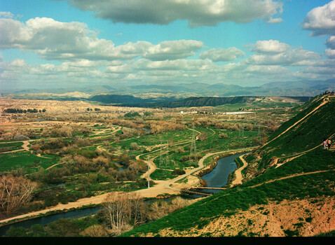 Aerial view of a winding river through cultivated valley with forested hillsides and distant mountains under partly cloudy sky.
