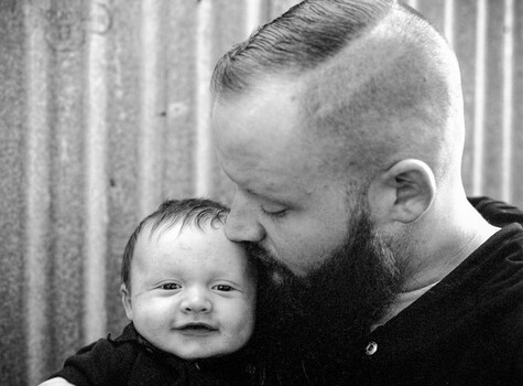 Bearded man in black shirt tenderly kissing forehead of smiling infant.