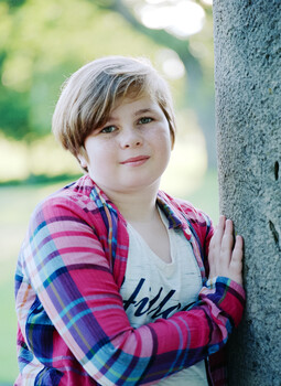 Young person in pink plaid shirt leaning against tree trunk outdoors.