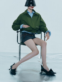 Person seated on metal chair wearing green jacket and dark shorts, posed with extended legs against white background.
