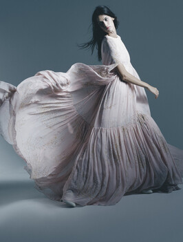 Woman in flowing white pleated dress against neutral blue-gray background.