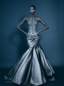 Model in metallic silver mermaid gown with structured bodice against blue backdrop.