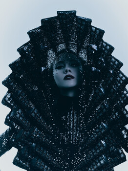 Portrait of a figure with an elaborate tiered collar made of sequined fabric, shot against a light background.