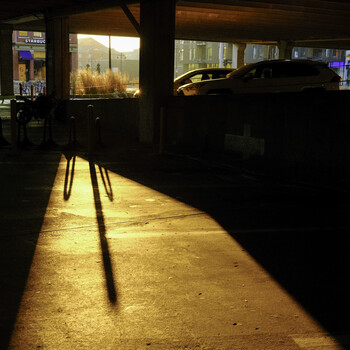Long shadows cast across a concrete storefront at night, with illuminated shop windows and parked vehicles visible.